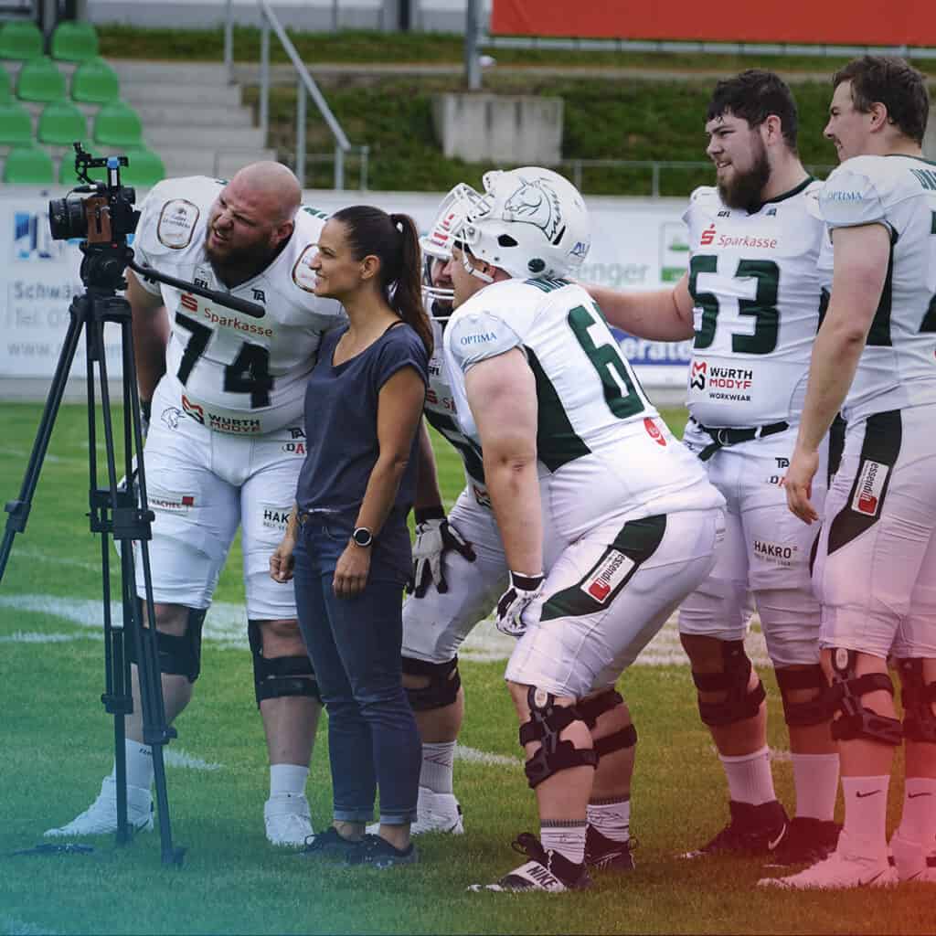 Yvonne Schulze umringt von Football-Spielern, die ihr kritisch über die Schulter schauen und das Kamerabild kontrollieren
